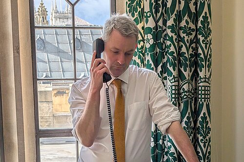 Will Forster speaking on the phone in his Westminster office, he is wearing a white shirt with the sleeves rolled up and a yellow tie.
