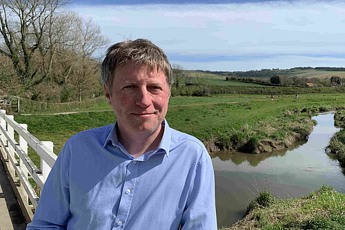 James MacCleary, in the foreground, on the South Downs Way near Alfriston, with a stream, fields, trees and blue sky behind him