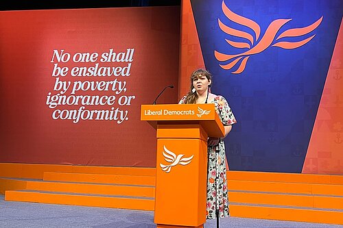 Katharine Macy at the Conference podium in a hall with bold coloured panels and a Lib Dem logo behind her