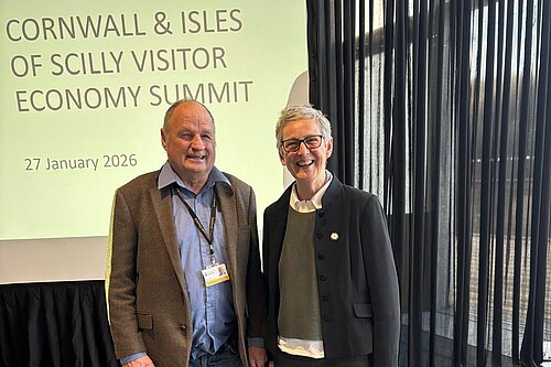 Cornwall councillors Jim Candy and Sarah Preece stand in front of a slide projection. The slide text reads: Cornwall & Isles Of Scilly Visitor Economy Summit. 