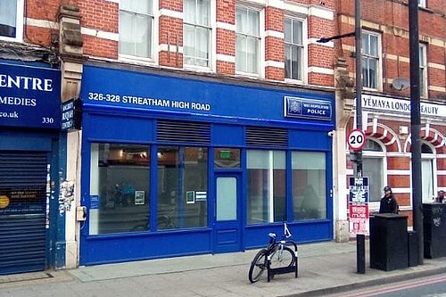 Photo of a road and pavement in the foreground then a line of shops, one of which is a police station
