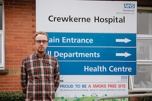 Adam Dance MP standing in front of a sign for Crewkerne Hospital in Somerset.