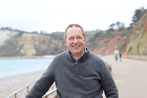 A photo of Richard Foord on the seafront at Seaton. The background is out of focus, with the rest of the promenade and cliffs in the background. There are also a couple of people visible in the distance. The weather appears overcast.