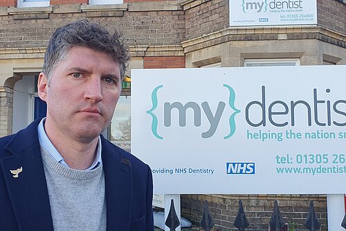 Lib Dem Candidate Edward Morello standing in front of an NHS dentist sign.